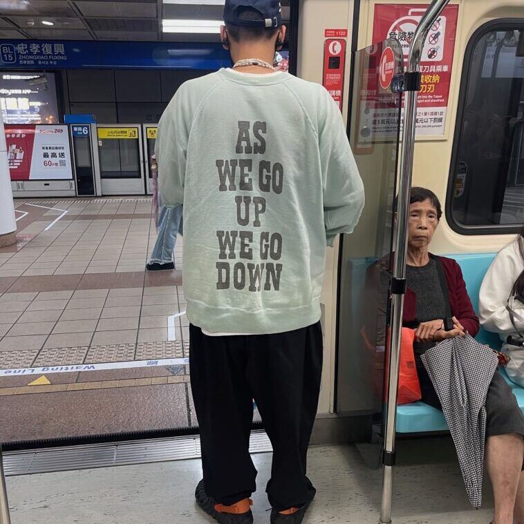 young man in a giant sweater. On the back, in large letters, it says 'as we go up we go down'